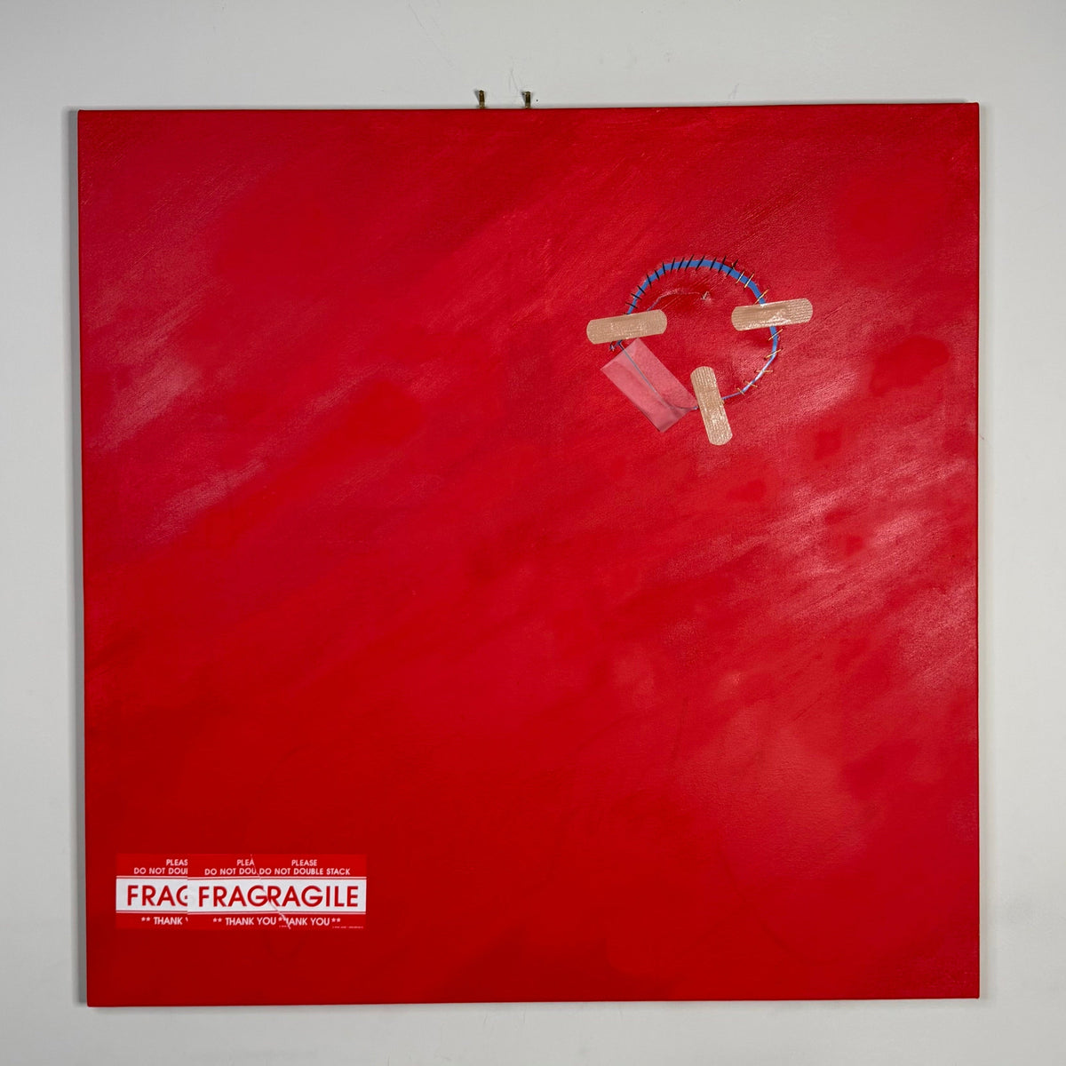 A square red wall art piece hanging on a light gray wall, featuring a small white label reading 'FRAG FRAGAGILE' near the bottom left and a small circular arrangement of wooden blocks and blue string near the upper center.