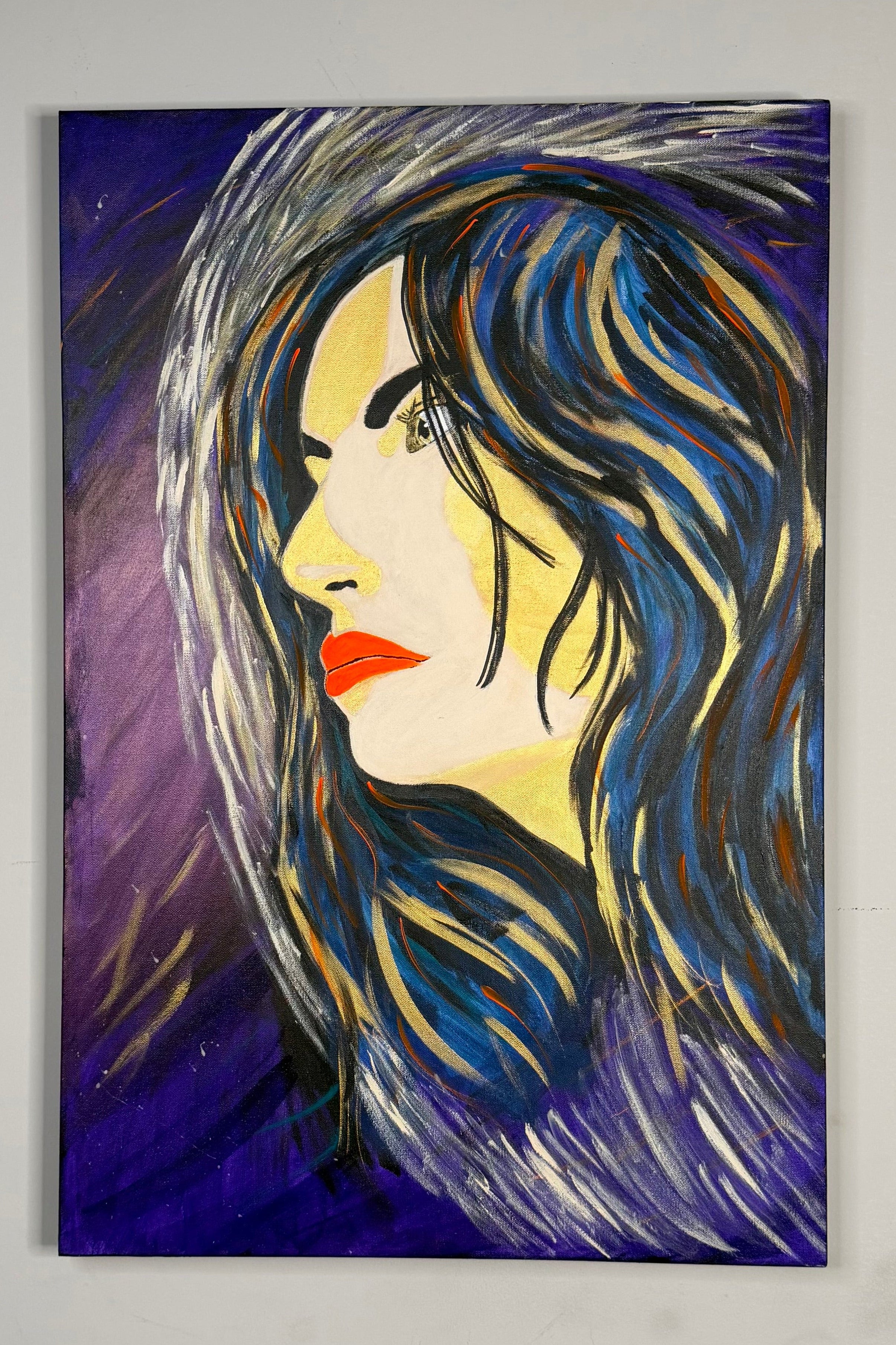 A vertical canvas painting of a woman's profile with flowing dark blue and black hair, yellow skin tones, and red lips against a deep purple background, displayed on a light gray wall.